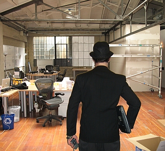 An ergonomist enters an office under construction, holding laptop & recorder